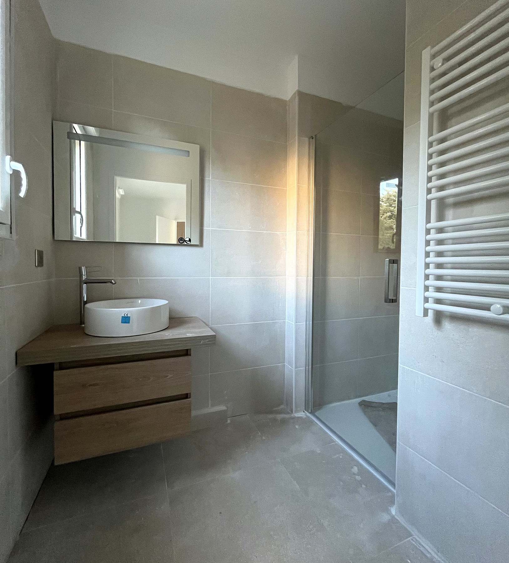 La même salle de bain vue depuis l’entrée : sèche-serviettes à droite, douche à l’italienne à receveur extra-plat au fond, plan de toilette à gauche sous le miroir.