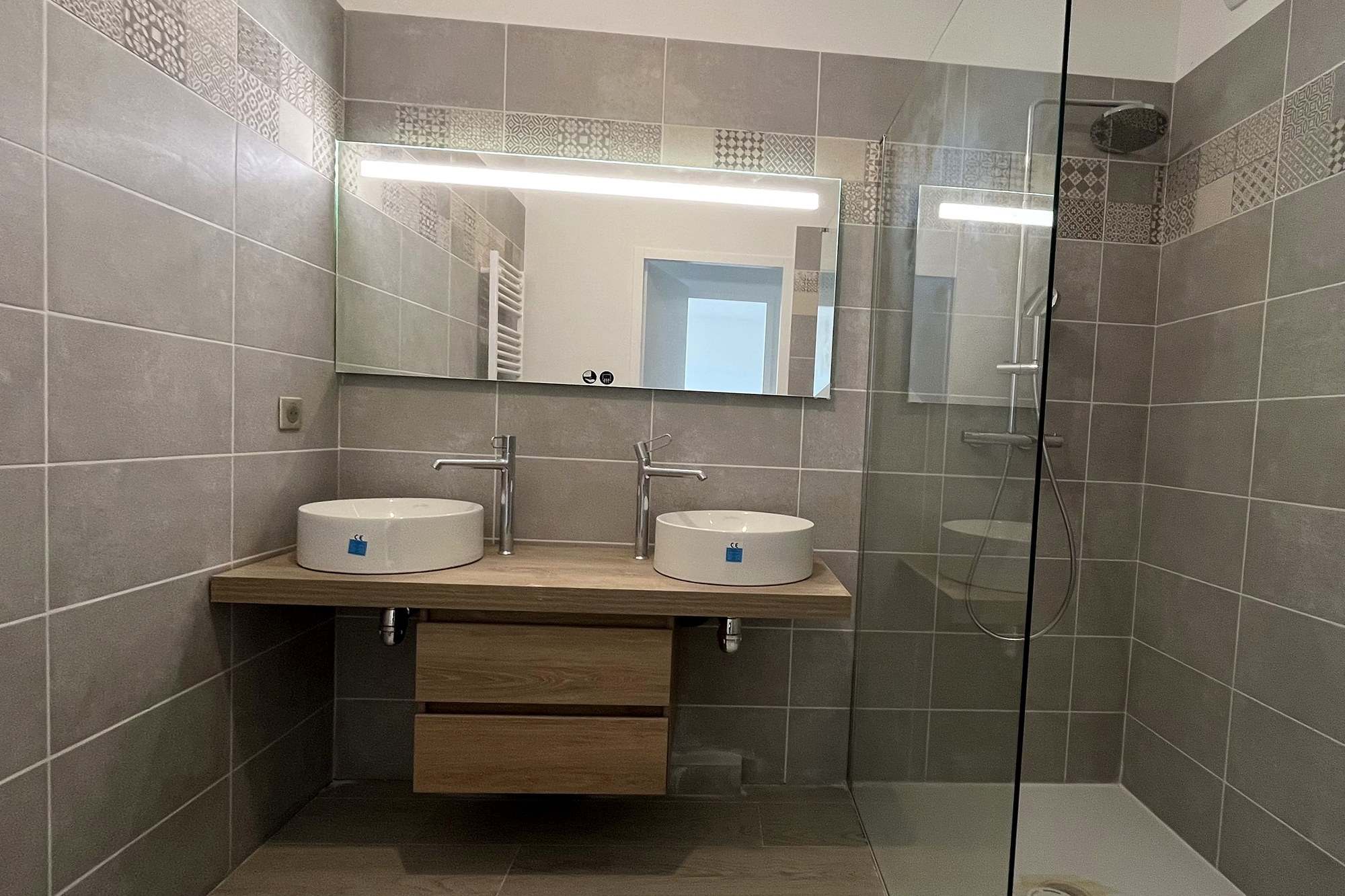 Salle de bain terminée : double vasque posée sur un plan en bois, grand miroir éclairé, douche à l’italienne fermée par une paroi vitrée, carrelage grand format effet pierre.