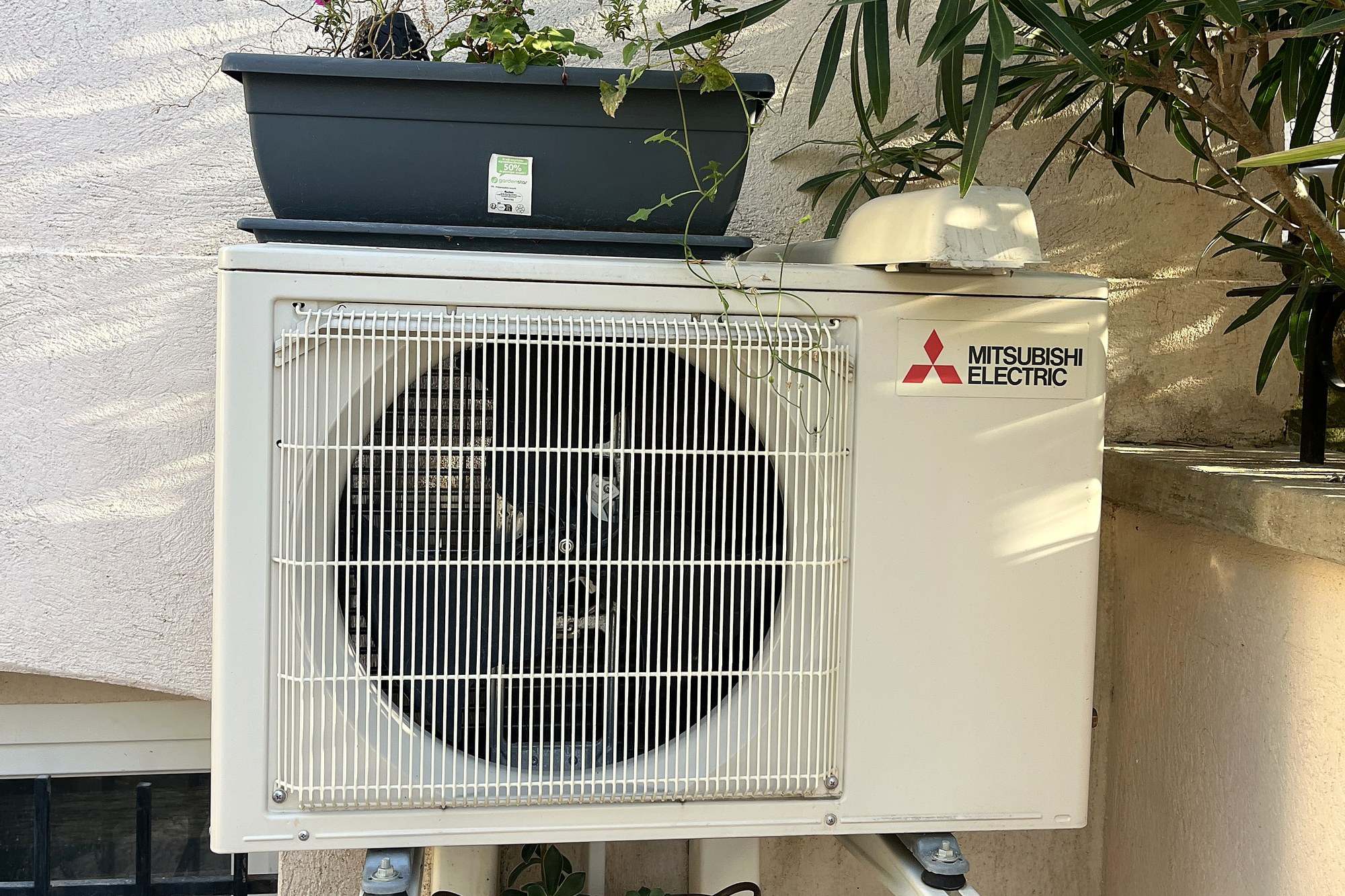 Deux groupes extérieurs Mitsubishi Electric superposés sur consoles murales, au pied d’un laurier-rose, une jardinière fleurie posée sur celui du haut.