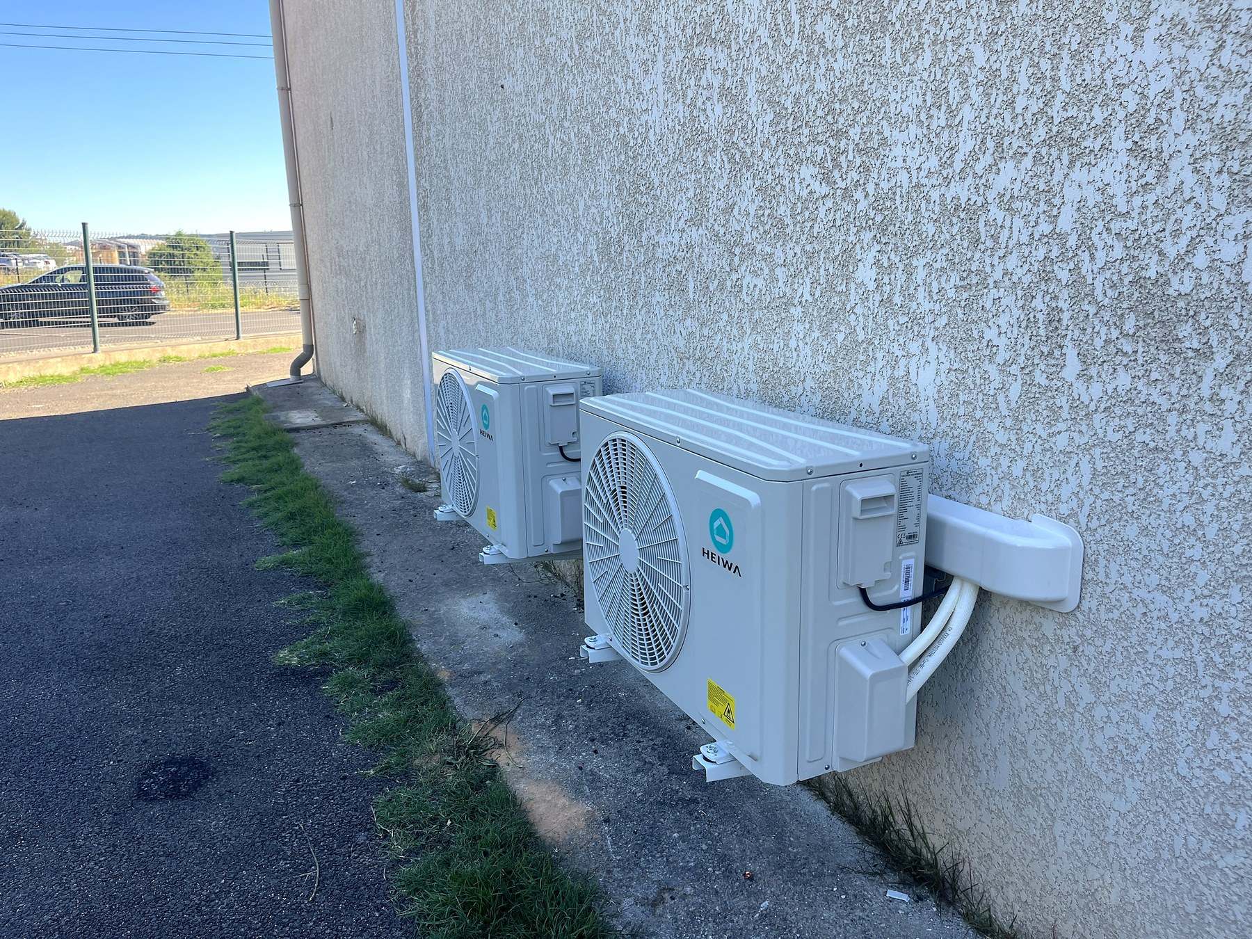 Deux groupes extérieurs Heiwa fixés sur consoles murales le long d’une façade en crépi, leurs liaisons entrant dans le mur par une goulotte courte.