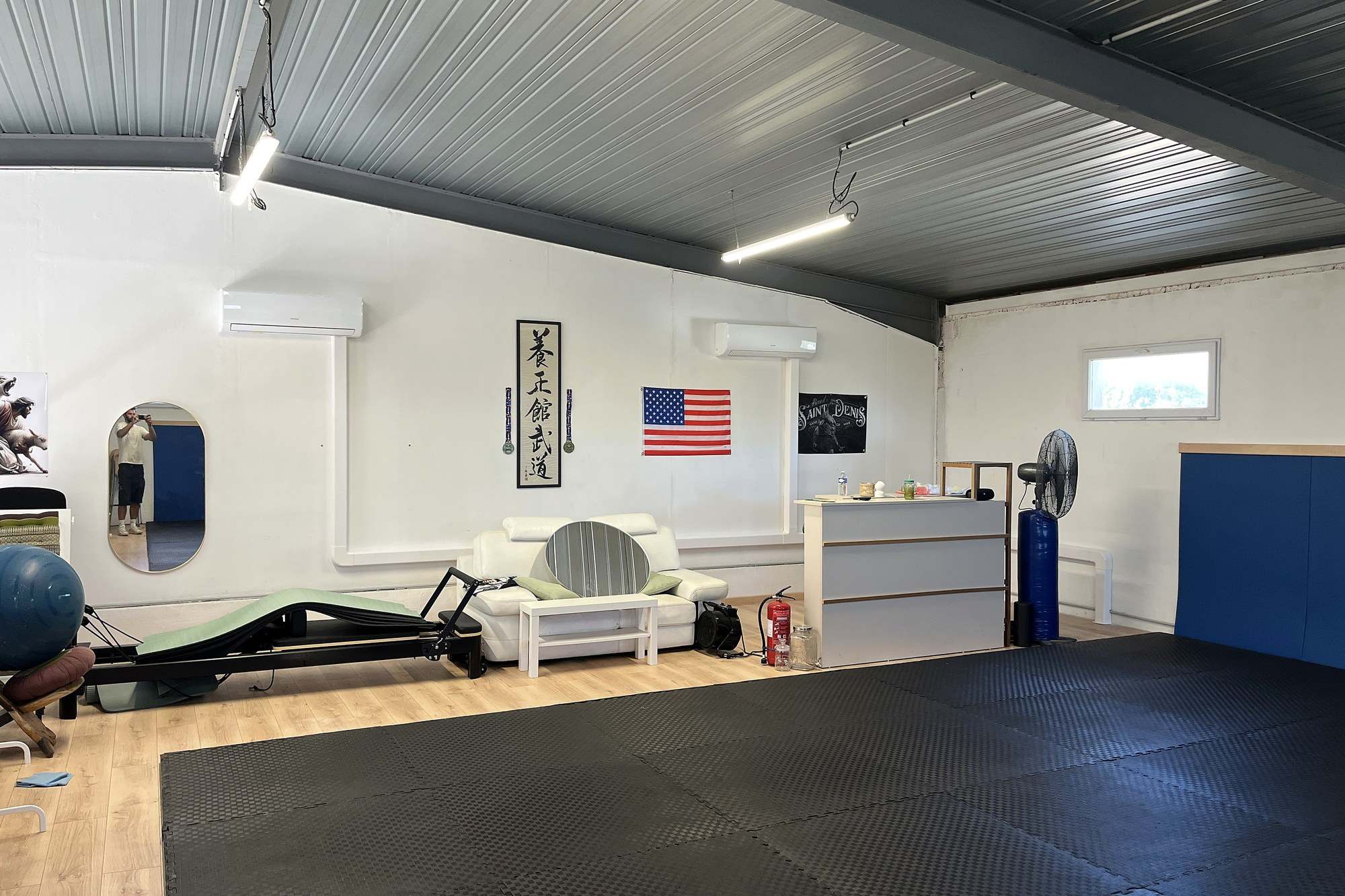 Salle de sport avec tatami noir et parquet clair, deux unités murales posées en haut des murs blancs sous une charpente métallique grise.