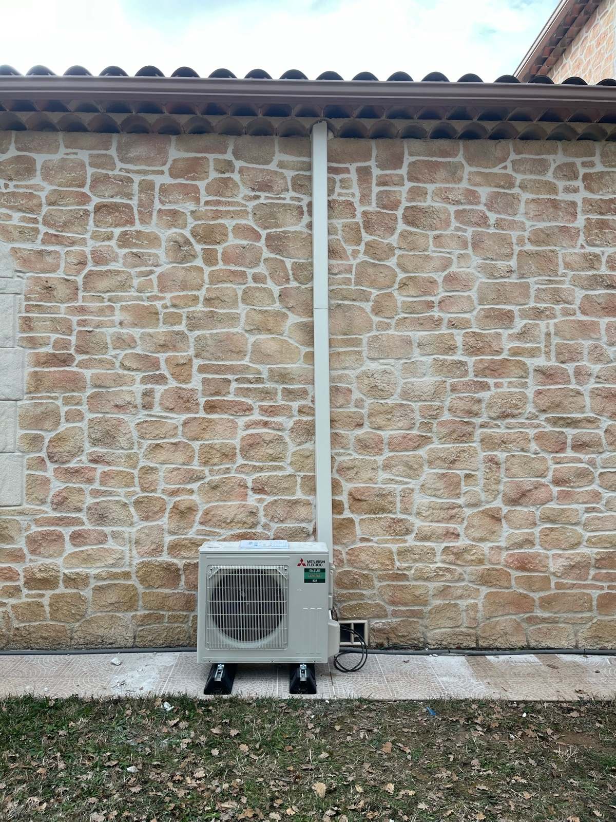 Groupe extérieur Mitsubishi Electric posé sur deux plots au pied d’un mur en pierre, sa goulotte blanche montant verticalement jusqu’à la génoise du toit.