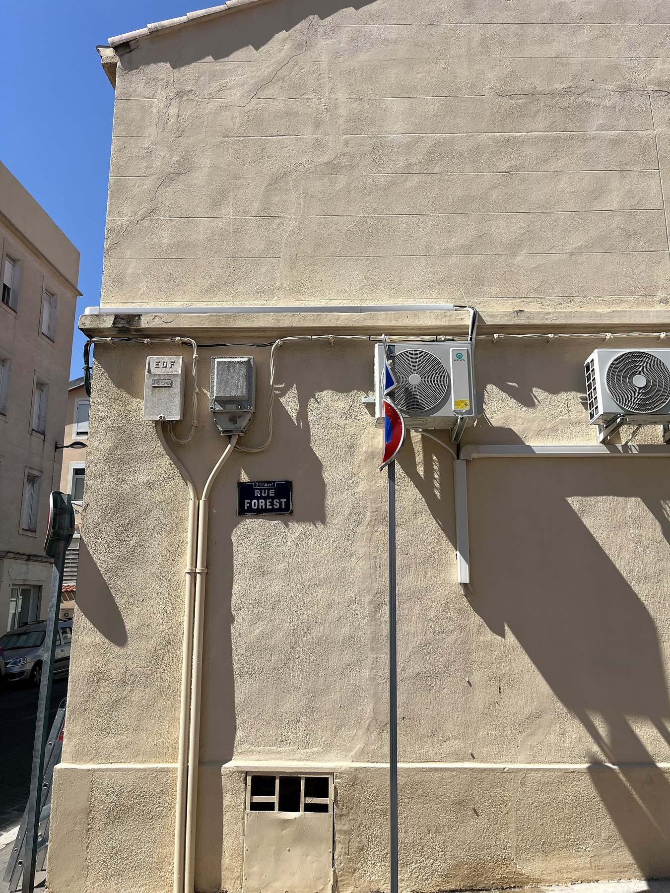 Angle de rue marseillais : deux groupes extérieurs fixés sur consoles en haut d’une façade beige, leurs liaisons cheminant sous la corniche jusqu’au coffret, plaque de rue du 7ᵉ arrondissement visible en dessous.