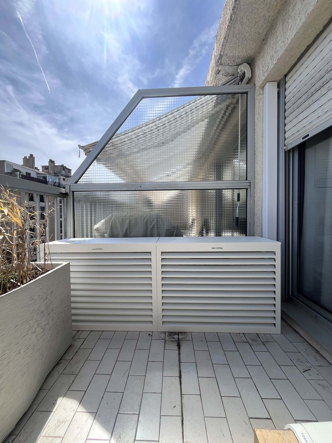 Deux caches techniques à lames blanches alignés sur une terrasse carrelée d’immeuble, devant une verrière, avec les toits marseillais en arrière-plan.