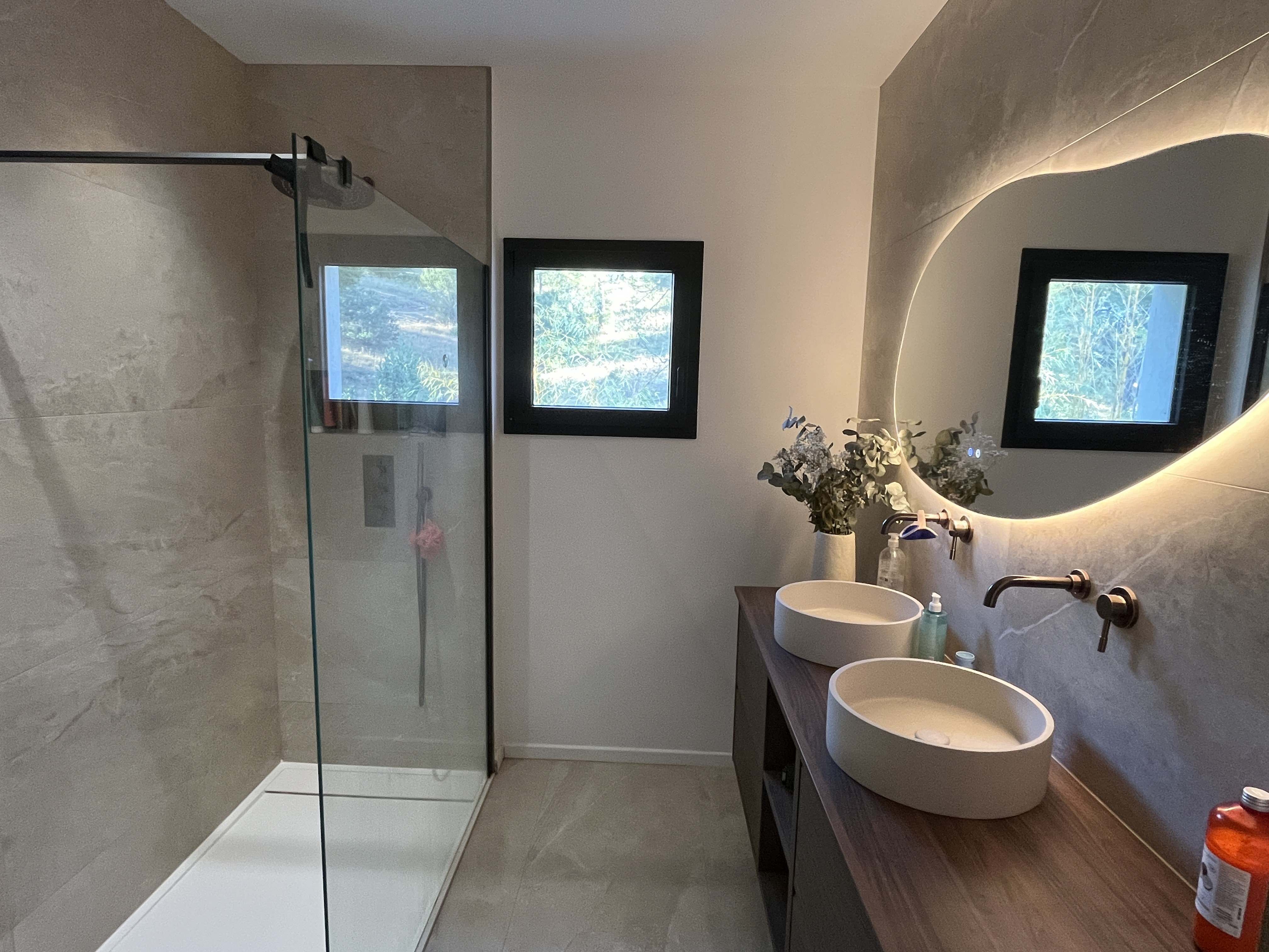 La même salle de bain vue depuis l’entrée : la paroi de douche au premier plan, le plan de toilette et ses deux vasques au fond à droite sous le miroir rétroéclairé.