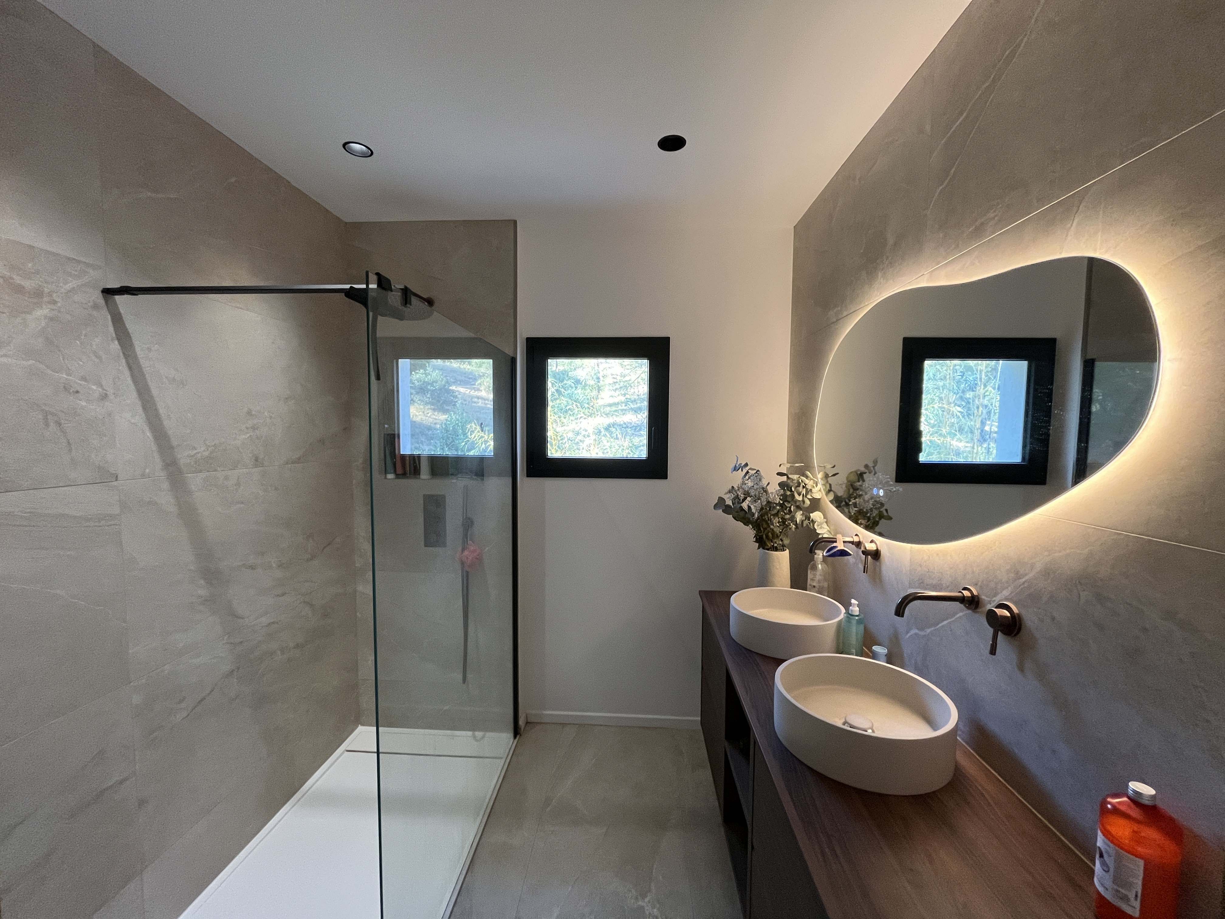 Salle de bain terminée vue en enfilade : douche à l’italienne à receveur extra-plat fermée par une paroi vitrée fixe, deux fenêtres à menuiserie noire, miroir organique rétroéclairé, deux vasques ovales sur un plan en bois et carrelage grand format effet pierre au mur comme au sol.