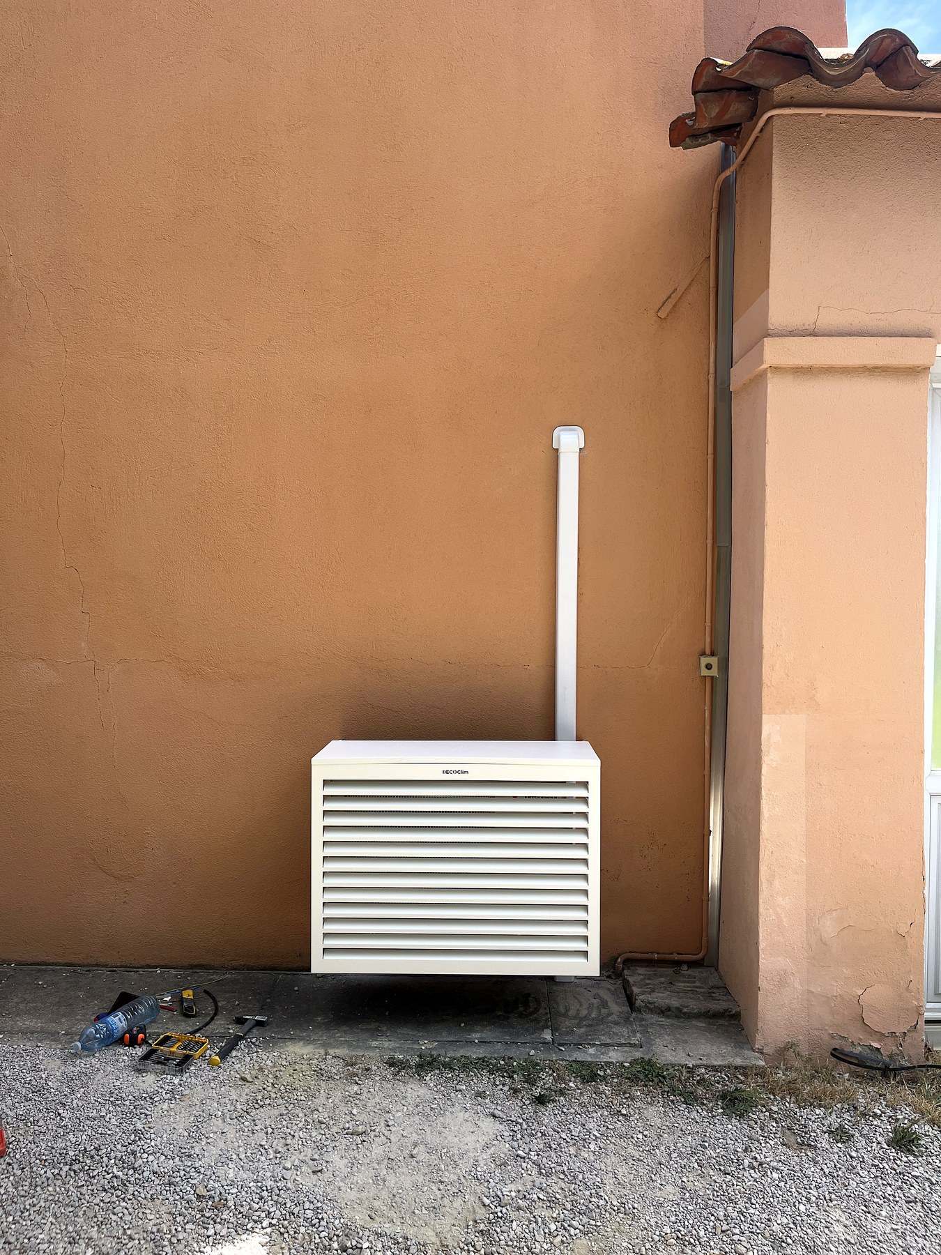 Cache technique à lames horizontales dissimulant le groupe extérieur au pied d’un mur enduit ocre, la ligne des condensats montant verticalement le long de l’angle.
