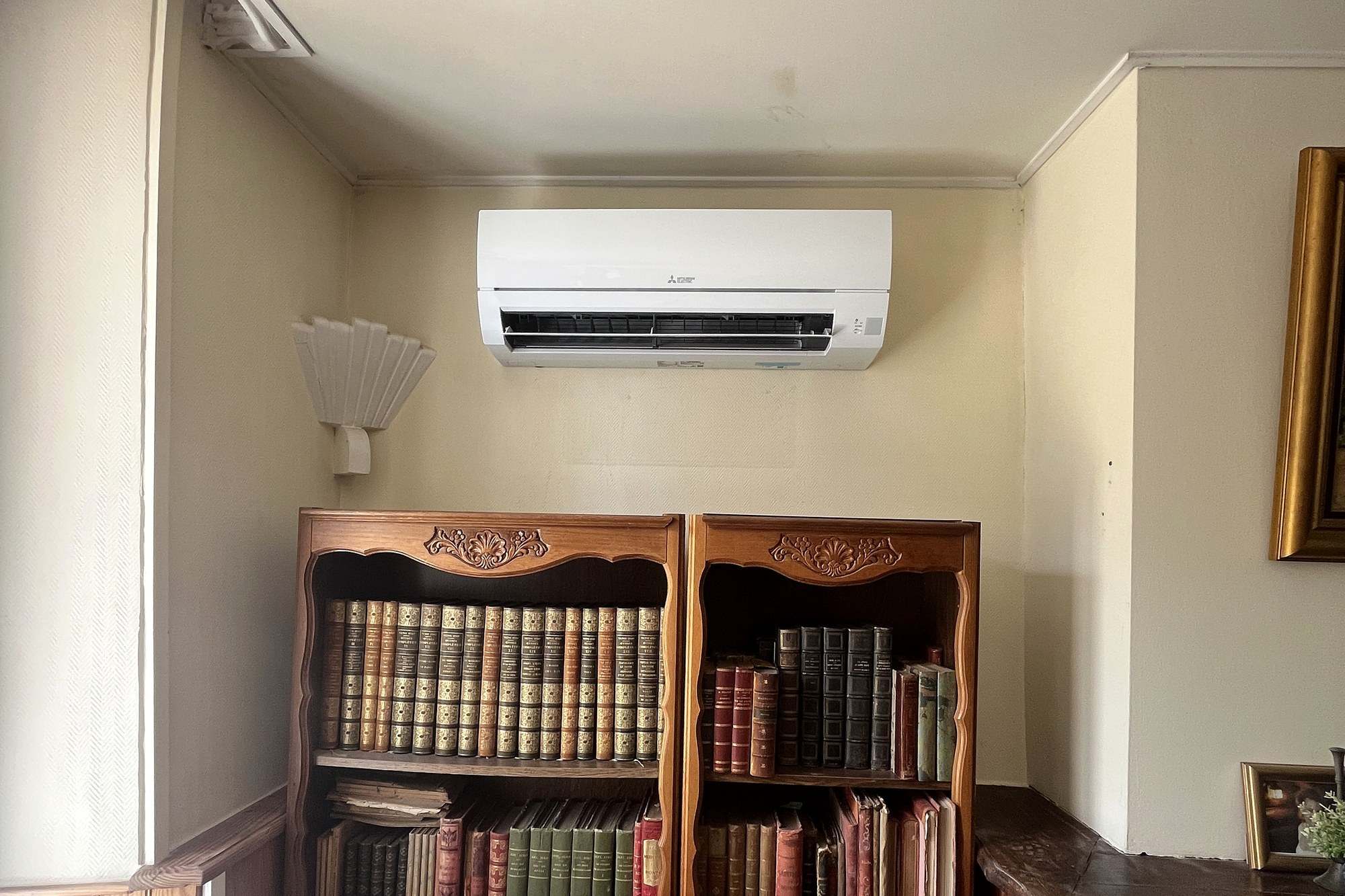 Unité murale Mitsubishi Electric posée en haut d’un mur beige, au-dessus d’une bibliothèque en chêne garnie de livres anciens, à côté d’une cheminée en brique.