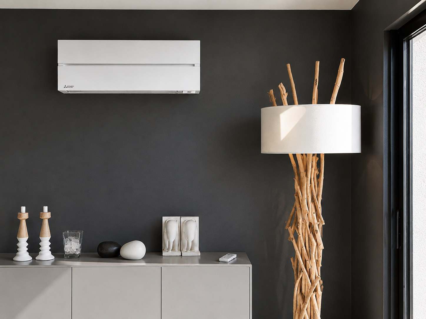 Unité murale Mitsubishi Electric posée en haut d’un mur anthracite dans un séjour contemporain, au-dessus d’un buffet blanc, à côté d’un lampadaire en bois flotté.